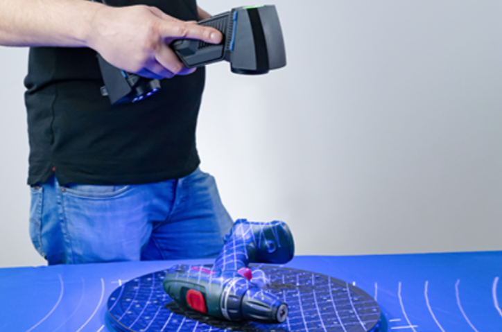 Rankinis 3D skeneris su mėlynu apšvietimu skenuoja automobilio padangą, šalia matomas nuskenuotas protektoriaus vaizdas ekrane.