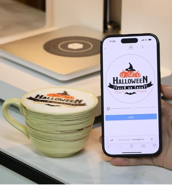 Išmanusis telefonas su atidaryta kavos spausdinimo programa ir šalia esantis puodelis su personalizuotu Halloween dizainu.