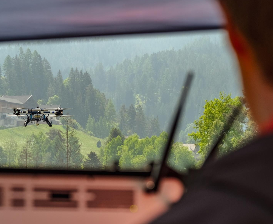 FB3 krovininis dronas skrenda virš kalnų slėnio, vaizdas pro automobilio priekinį stiklą.