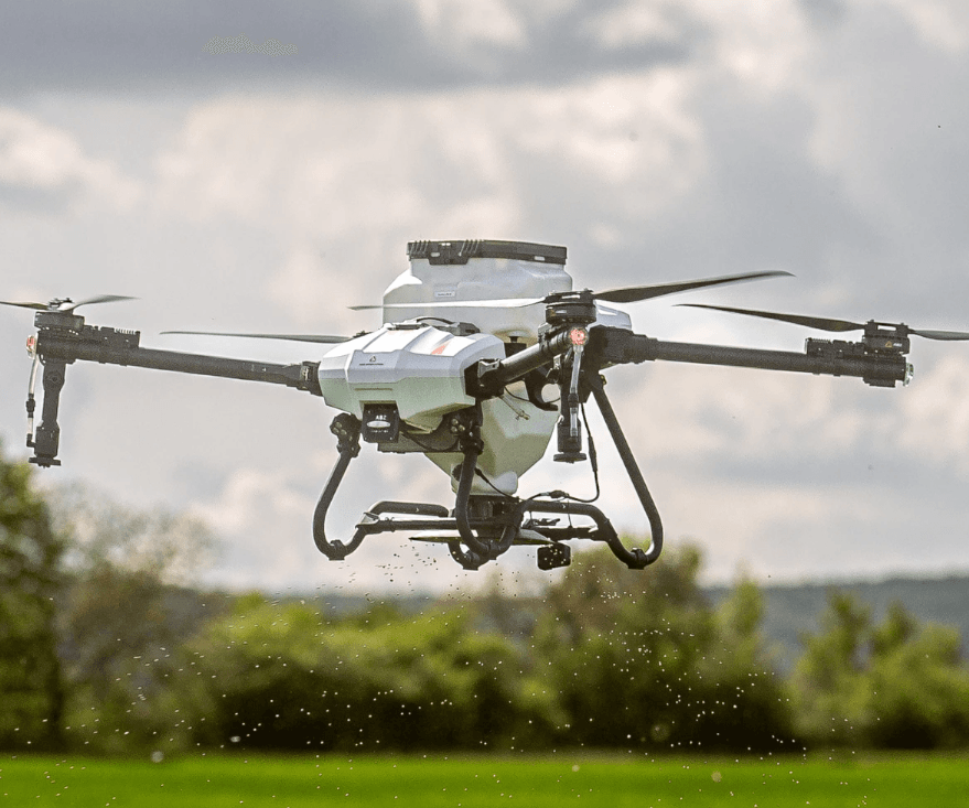 Purškimo dronas skrydyje virš lauko, skirtas augalų priežiūrai ir vienodam purškimo padengimui realiomis sąlygomis.