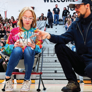 Instruktorius padeda mokinei valdyti droną varžybų metu sporto salėje, fone matyti žiūrovai.