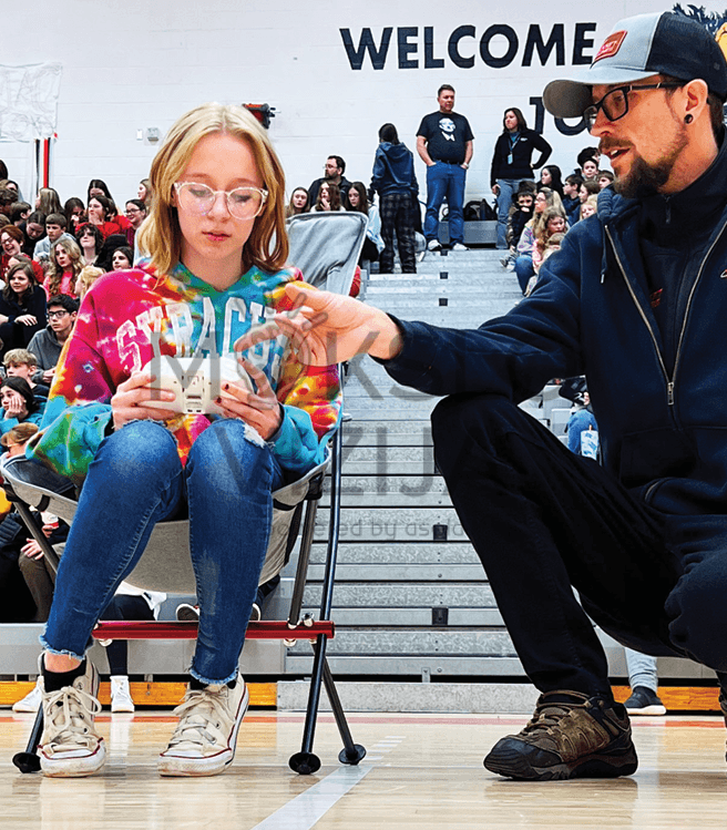 Instruktorius padeda mokinei valdyti droną varžybų metu sporto salėje, fone matyti žiūrovai.