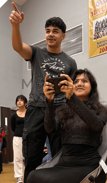 Mokiniai sporto salėje valdo droną nuotolinio valdymo pultu, vyksta praktinis mokymas ir treniruotė.