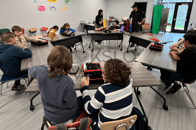 Mokiniai klasėje mokosi dronų valdymo, prie stalų matyti dronų dėklai ir mokymo priemonės.