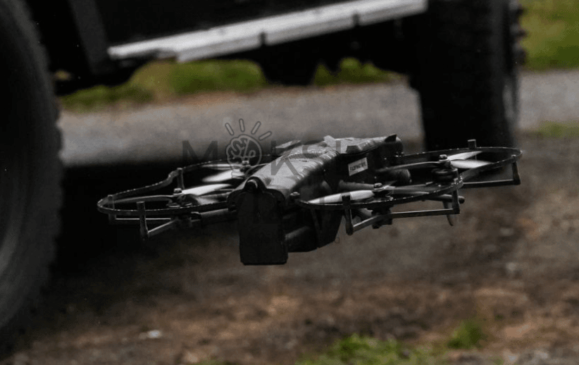 Taktinis dronas su apsaugotais rotoriais skrenda žemai virš žemės lauko sąlygomis prie transporto priemonės.