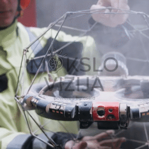 Operatorius laiko inspekcinį droną su apsauginiu sferiniu rėmu, matoma pramoninė aplinka ir dūmai fone.