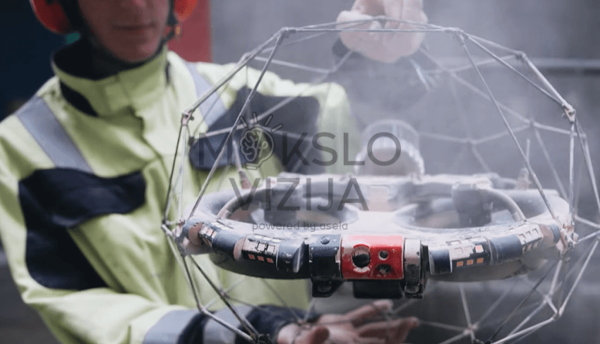 Operatorius laiko inspekcinį droną su apsauginiu sferiniu rėmu, matoma pramoninė aplinka ir dūmai fone.