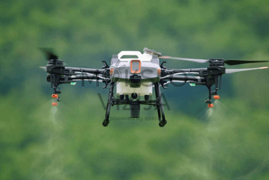 Žemės ūkio purškimo dronas purškia augalus skrydžio metu, matyti purškimo srovės ir žalios gamtos fonas.