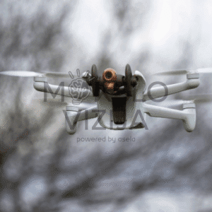 Baltas kompaktiškas dronas su integruota kamera skrenda ore, fone matyti neryškūs medžių siluetai.