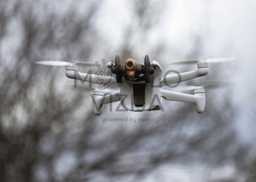 Baltas kompaktiškas dronas su integruota kamera skrenda ore, fone matyti neryškūs medžių siluetai.