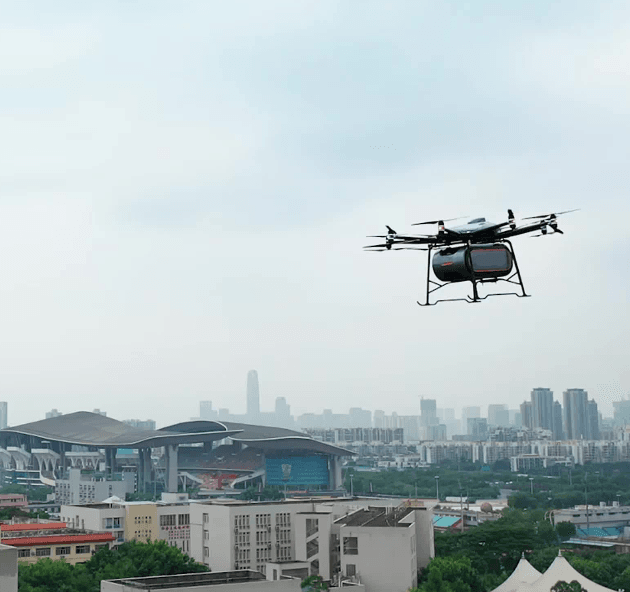 Krovininis dronas skrenda virš miesto, fone matyti pastatai ir stadionas, dronas gabena krovinio modulį.