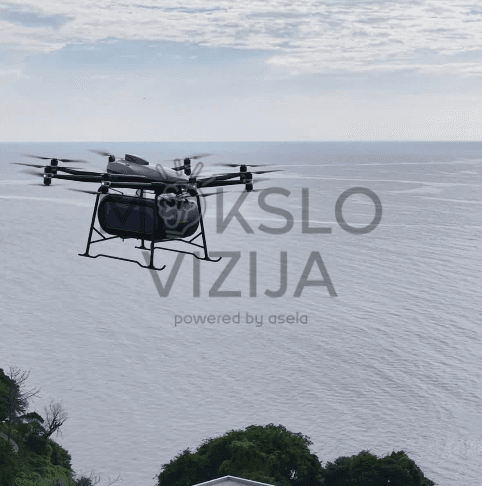 Krovininis dronas skrenda virš jūros pakrantės, fone matyti vanduo ir horizontas, dronas gabena krovinį.