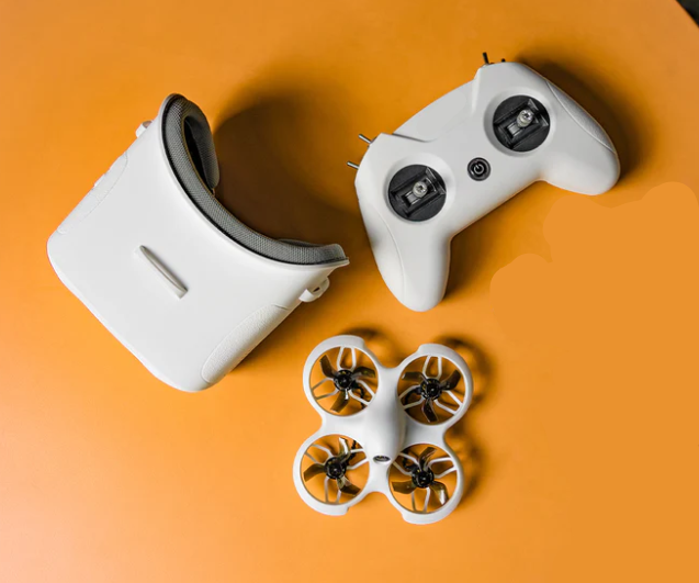 FPV drono komplektas ant oranžinio fono – akiniai, nuotolinio valdymo pultas ir mini dronas su apsaugotais propeleriais.
