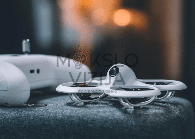 Baltas mini FPV dronas su integruota kamera padėtas ant audinio paviršiaus, šalia matomas valdymo pultas.