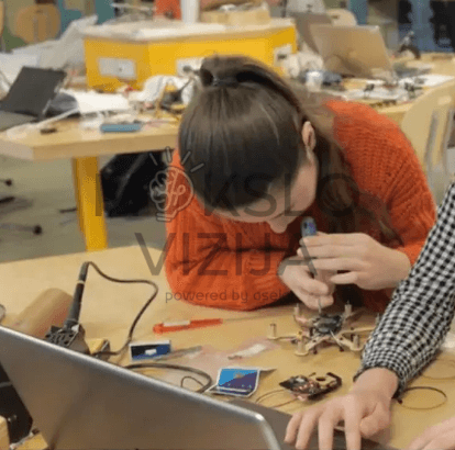 Mokiniai prie stalo konstruoja elektronikos ir drono komponentus, matomas litavimo įrankis, detalės ir kompiuteris.