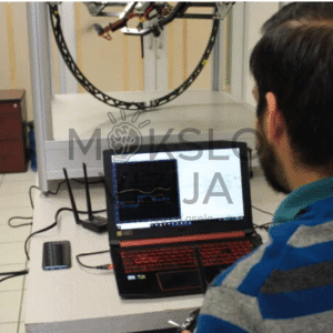 Operatorius sėdi prie nešiojamo kompiuterio ir stebi skrydžio duomenų grafiką, fone matomas giroskopinis dronų testavimo stendas.