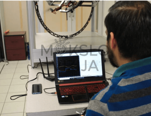 Operatorius sėdi prie nešiojamo kompiuterio ir stebi skrydžio duomenų grafiką, fone matomas giroskopinis dronų testavimo stendas.