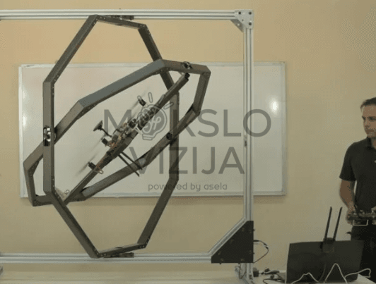 Dronas pritvirtintas giroskopiniame stende, matomas pasvirimo kampo testavimas ir valdymo pultas šalia.