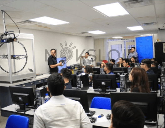 Mokymo klasė su studentais, dėstytojas pristato drono testavimo stendą ir valdymo sprendimus prie darbo vietų su kompiuteriais.
