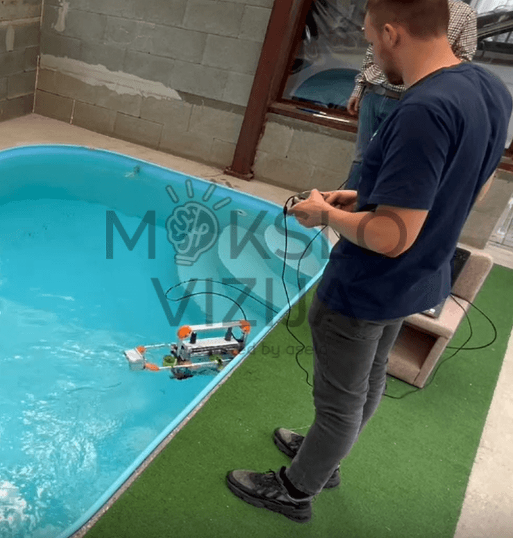 Operatorius valdo povandeninį robotą baseine, matomas kabelinis ryšys ir roboto judėjimas vandenyje.