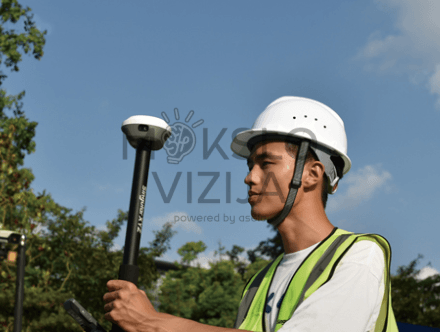 Operatorius su apsauginiu šalmu laiko RTK GNSS anteną ant stulpo ir ruošiasi atlikti matavimus lauko aplinkoje.