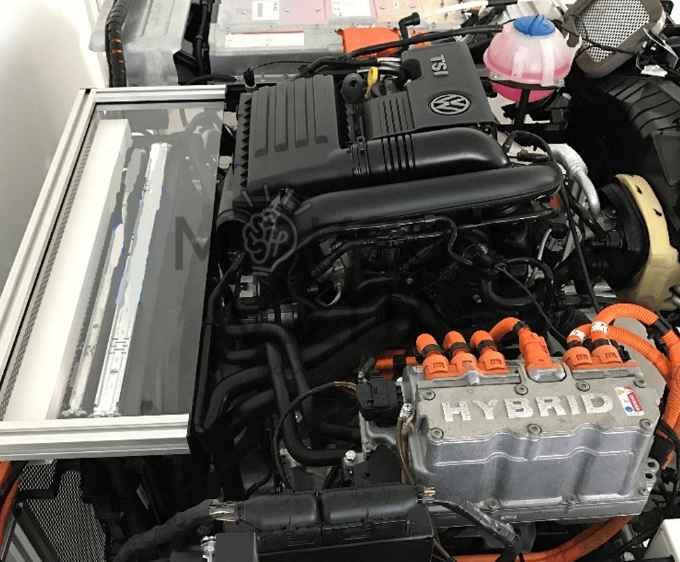 Hibridinės sistemos komponentų artimas vaizdas su „HYBRID“ moduliu, aukštos įtampos oranžiniais kabeliais, TSI variklio mazgu ir aušinimo skysčio bakeliu mokymo stende.
