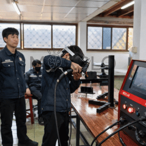 Mokinys atlieka suvirinimo pratimus virtualios realybės simuliatoriumi, šalia stebi kiti studentai mokymo dirbtuvėse.