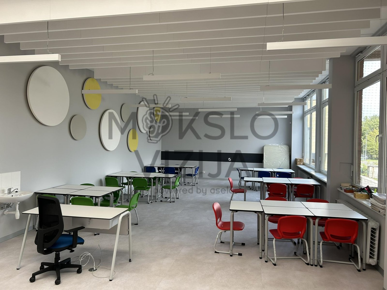 Moderni mokyklos klasė su mokinių stalais, spalvotomis kėdėmis ir akustinėmis sienų panelėmis.