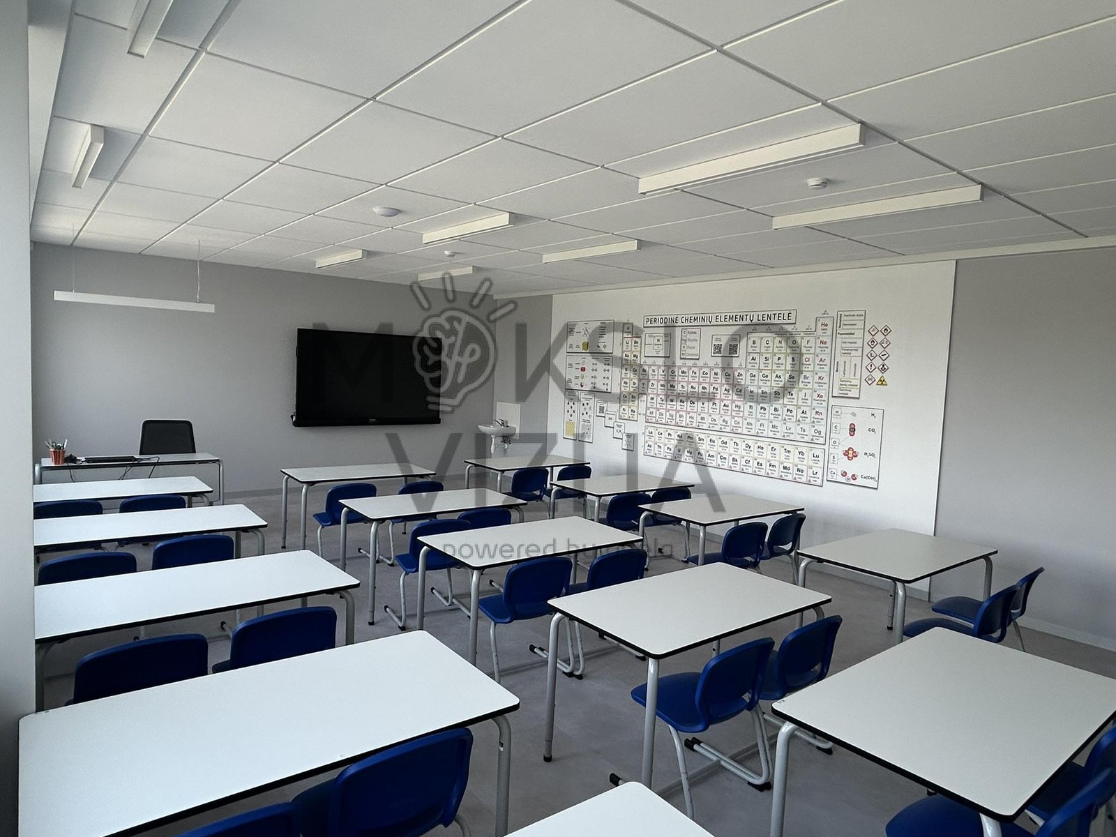 Moderni chemijos klasė su mokinių stalais, interaktyviu ekranu ir periodine elementų lentele ant sienos.