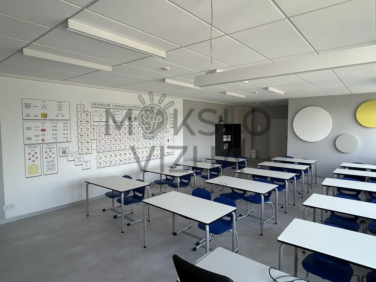 Moderni chemijos klasė su mokinių stalais ir periodine elementų lentele ant sienos.