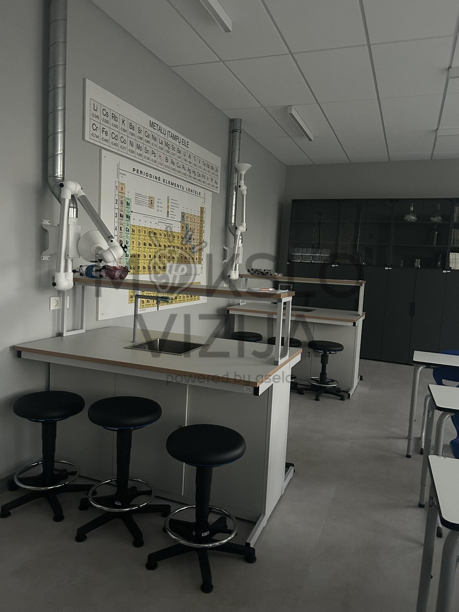 Moderni gamtos mokslų laboratorija su darbo stalais, ventiliacijos rankomis ir periodine elementų lentele.