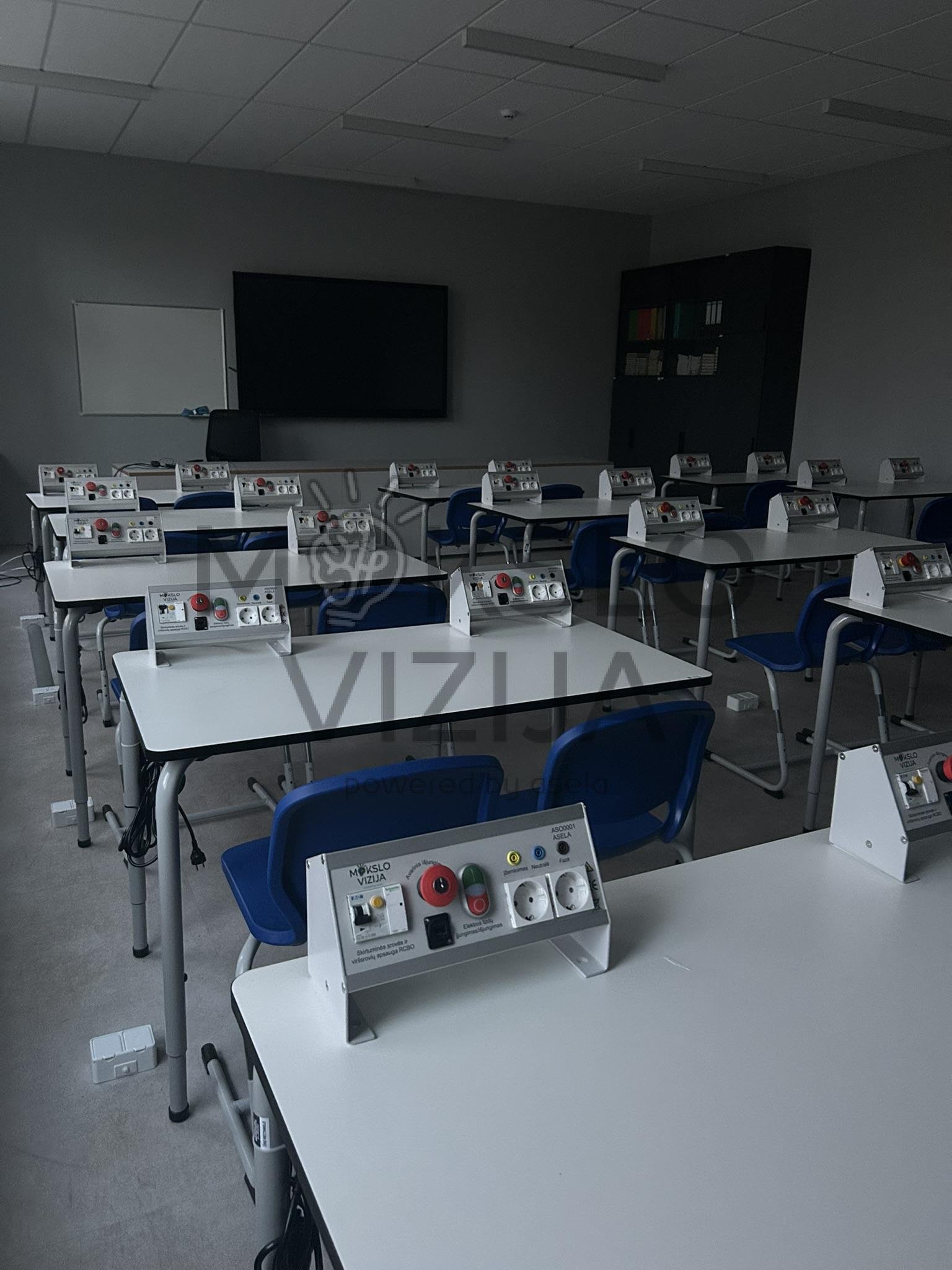 Fizikos klasė su mokinių stalais ir elektros eksperimentų valdymo moduliais ant stalų.
