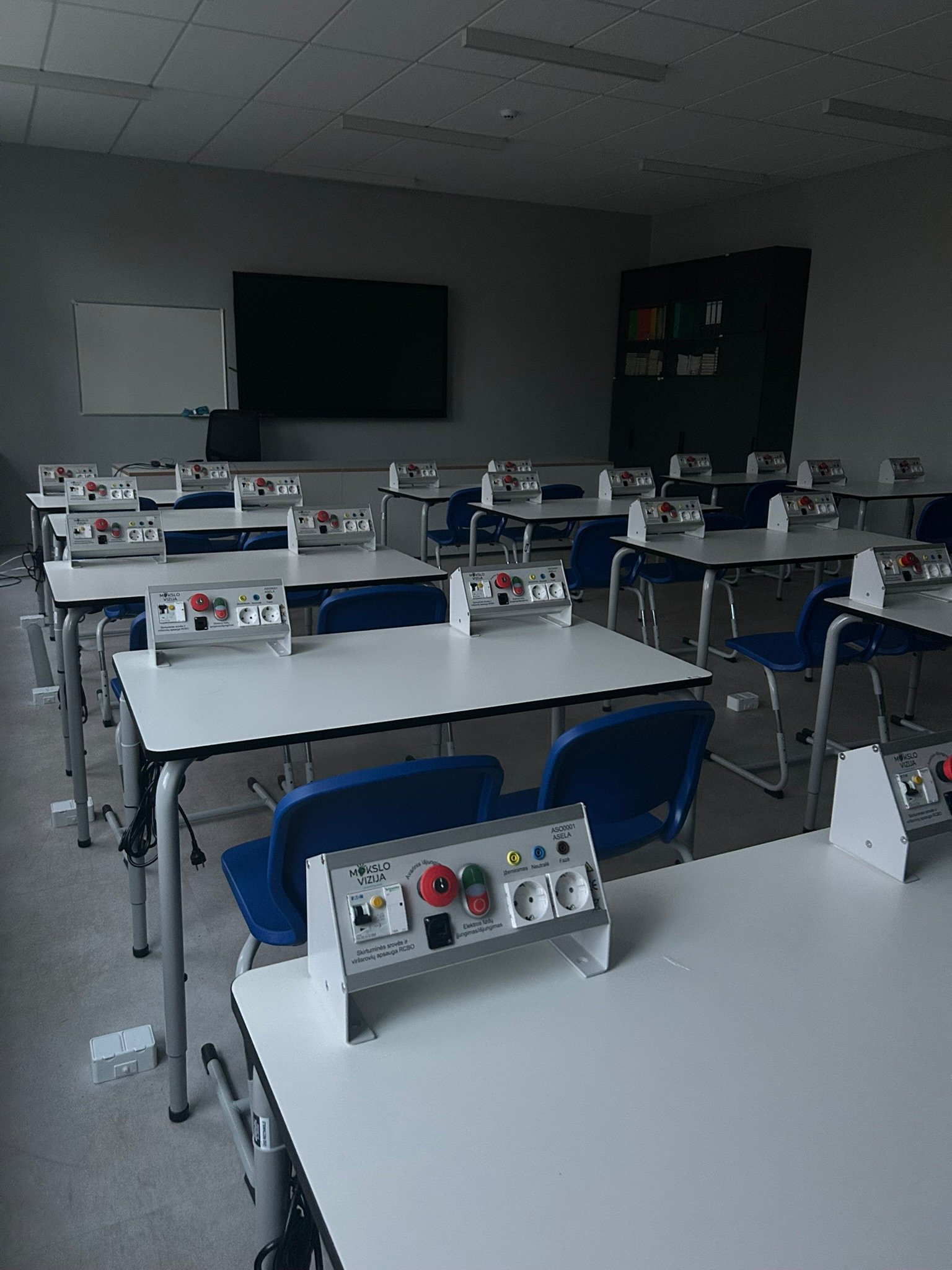 Fizikos klasė su mokinių stalais ir elektros eksperimentų valdymo moduliais ant stalų.