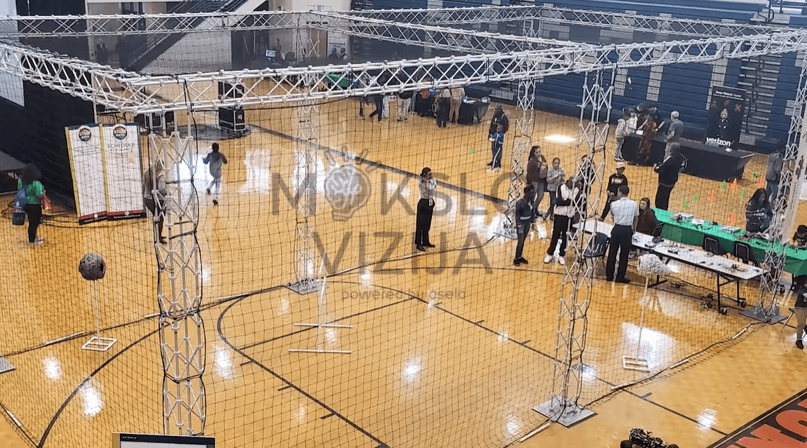 Varžybų dronų narvo konstrukcija sporto salėje su apsauginiu tinklu, skirta dronų mokymo ir varžybų organizavimui.