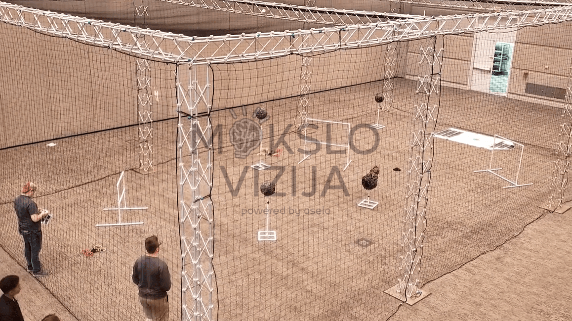 Didelis dronų skrydžių narvas su apsauginiu tinklu ir kliūčių elementais, naudojamas mokymuose patalpų viduje.