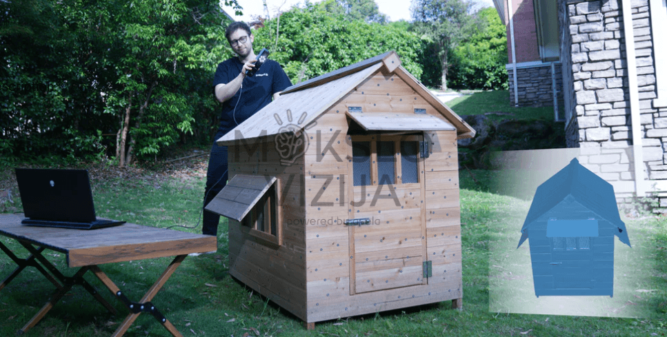 Žmogus lauke skenuoja medinį namelį nešiojamu 3D skeneriu, šalia prijungtas kompiuteris.