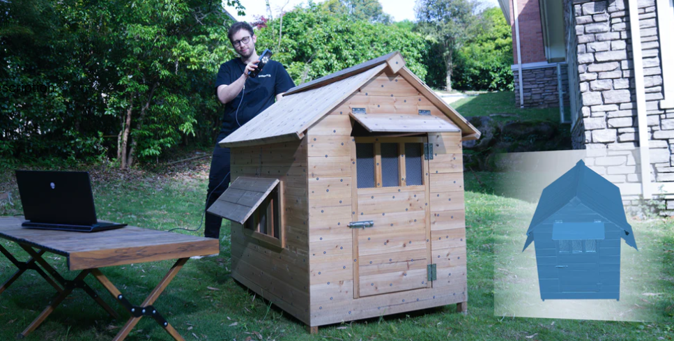 Žmogus lauke skenuoja medinį namelį nešiojamu 3D skeneriu, šalia prijungtas kompiuteris.