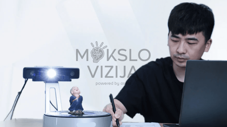 Stalinis 3D skeneris su ryškiu apšvietimu skenuoja objektą ant sukamo stalo, šalia dirba operatorius su kompiuteriu.