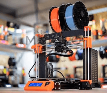 FDM 3D spausdintuvas su filamento ritėmis, pastatytas dirbtuvėse ant stalo, matomas spausdinimo mazgas.