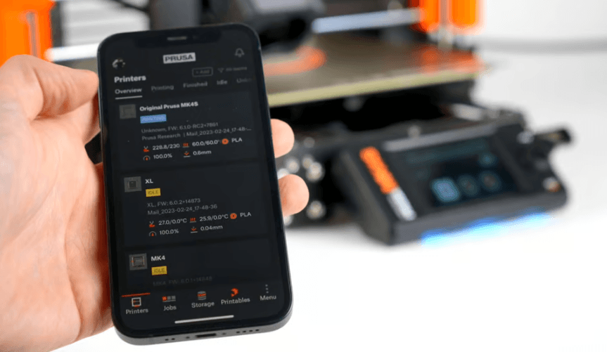 Rankoje laikomas mobilusis telefonas su Prusa programėle, kurioje matomas Original Prusa MK4S spausdintuvas ir jo spausdinimo būsena, fone matomas 3D spausdintuvas.