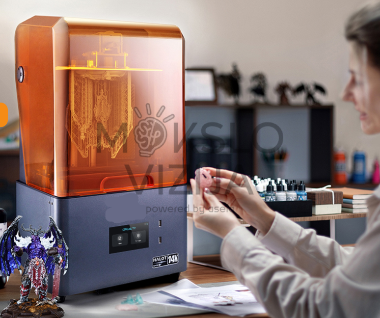 Creality HALOT-MAGE S 14K dervos 3D spausdintuvas su oranžiniu apsauginiu dangčiu, pastatytas ant stalo, šalia žmogus laiko mažą atspausdintą detalę rankose.