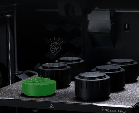Creality K2 Combo 3D spausdintuvo vidus, matoma spausdinimo platforma su keliais cilindriniais objektais ir vienu žalios spalvos modeliu.