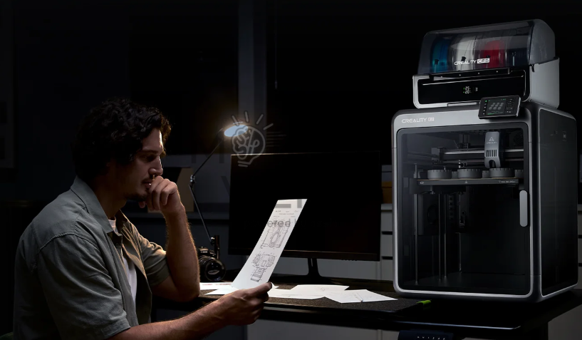 Žmogus sėdi prie darbo stalo ir laiko brėžinį, šalia stovi Creality K2 Combo 3D spausdintuvas su daugiaspalviu gijų moduliu ir apšviesta darbo zona.