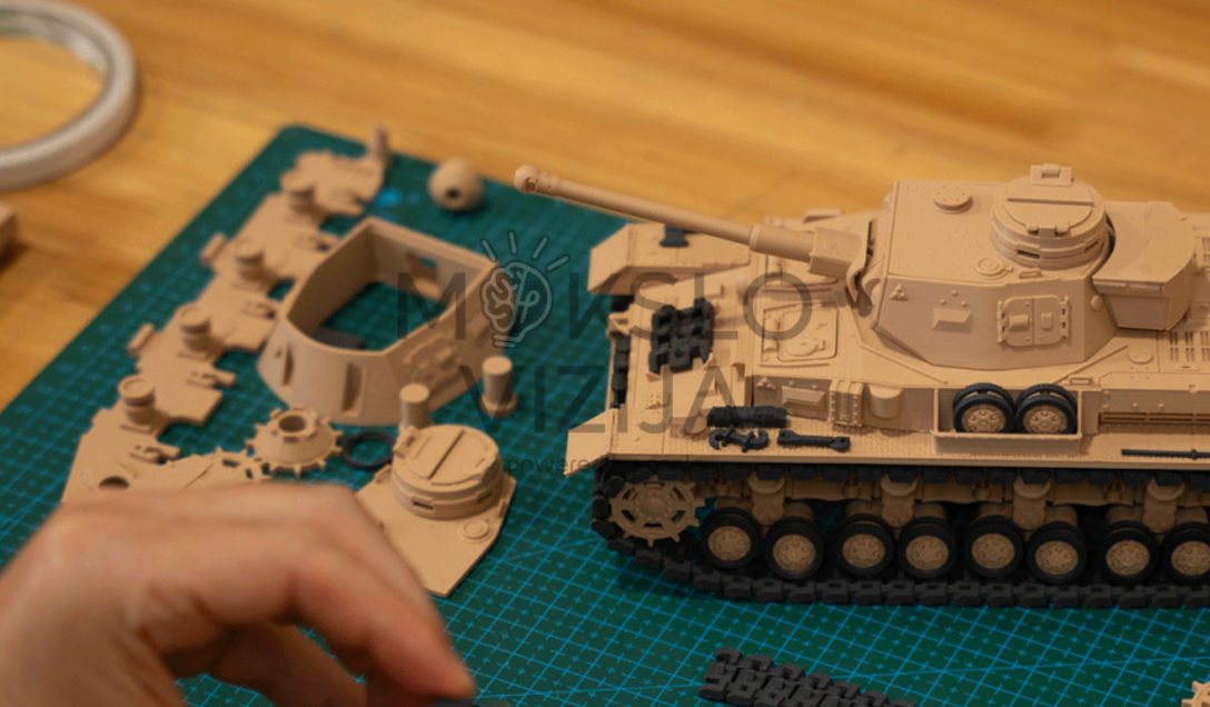 Ant žalio pjaustymo kilimėlio padėtos 3D spausdintos tanko modelio detalės ir surinktas miniatiūrinis tankas, matoma žmogaus ranka šalia.