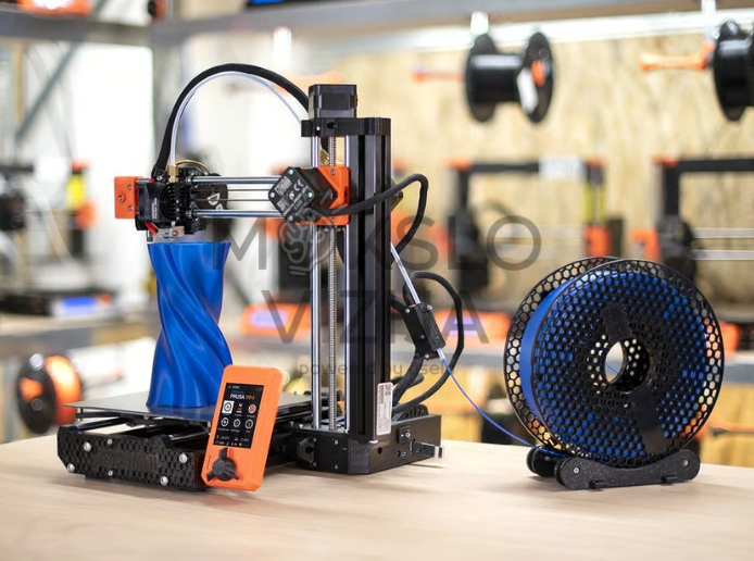 3D spausdintuvas ant stalo su atspausdintu mėlynu modeliu ant platformos, šalia padėta gijos ritė dirbtuvių aplinkoje.