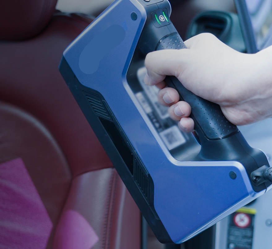 Rankoje laikomas mėlynos spalvos rankinis 3D skeneris, naudojamas skenuoti automobilio salono paviršių.