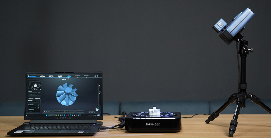 3D skenavimo sistema ant stalo, sudaryta iš nešiojamo kompiuterio su 3D modelio peržiūra, automatinės sukimo platformos ir skenerio, pritvirtinto prie trikojo.