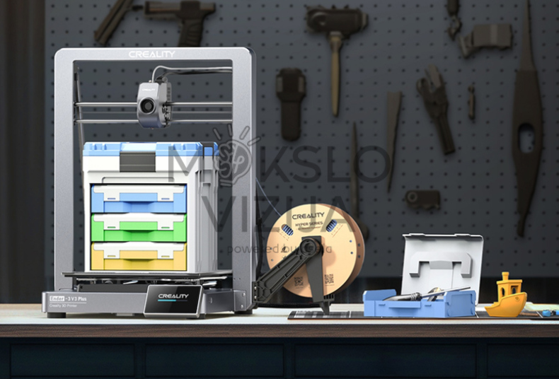 FDM tipo 3D spausdintuvas su metaliniu rėmu, dideliu spausdinimo tūriu, šalia padėta plastiko gijos ritė ir atspausdintos detalės ant darbo stalo dirbtuvėse.