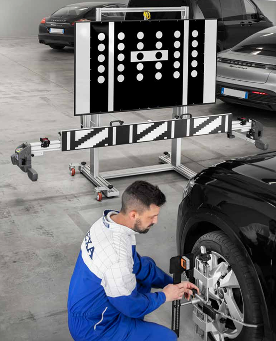 ADAS kalibravimo stendas su dideliu juodai baltu taikiniu automobilių servise, technikas reguliuoja automobilio jutiklius priekinėje dalyje.