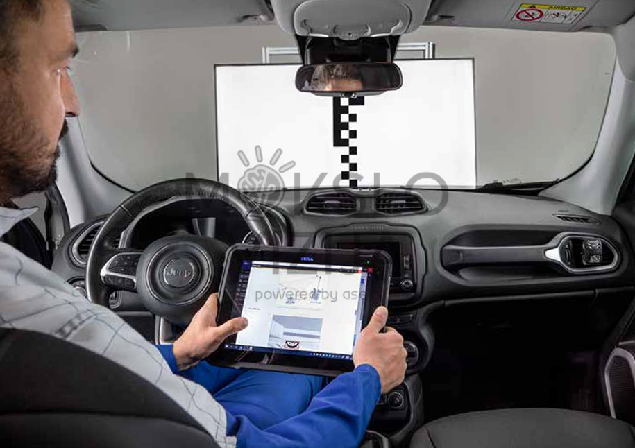 Automobilio salone sėdintis technikas su diagnostine planšete atlieka ADAS kameros kalibravimą priešais specialų kalibravimo ekraną.